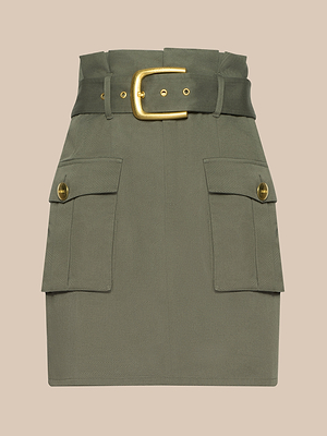 The Maelis Mini Skirt is an olive green cotton twill paperbag mini skirt with a wide matching belt and large gold buckle, featuring two front cargo pockets with gold buttons, shown against a plain beige background.