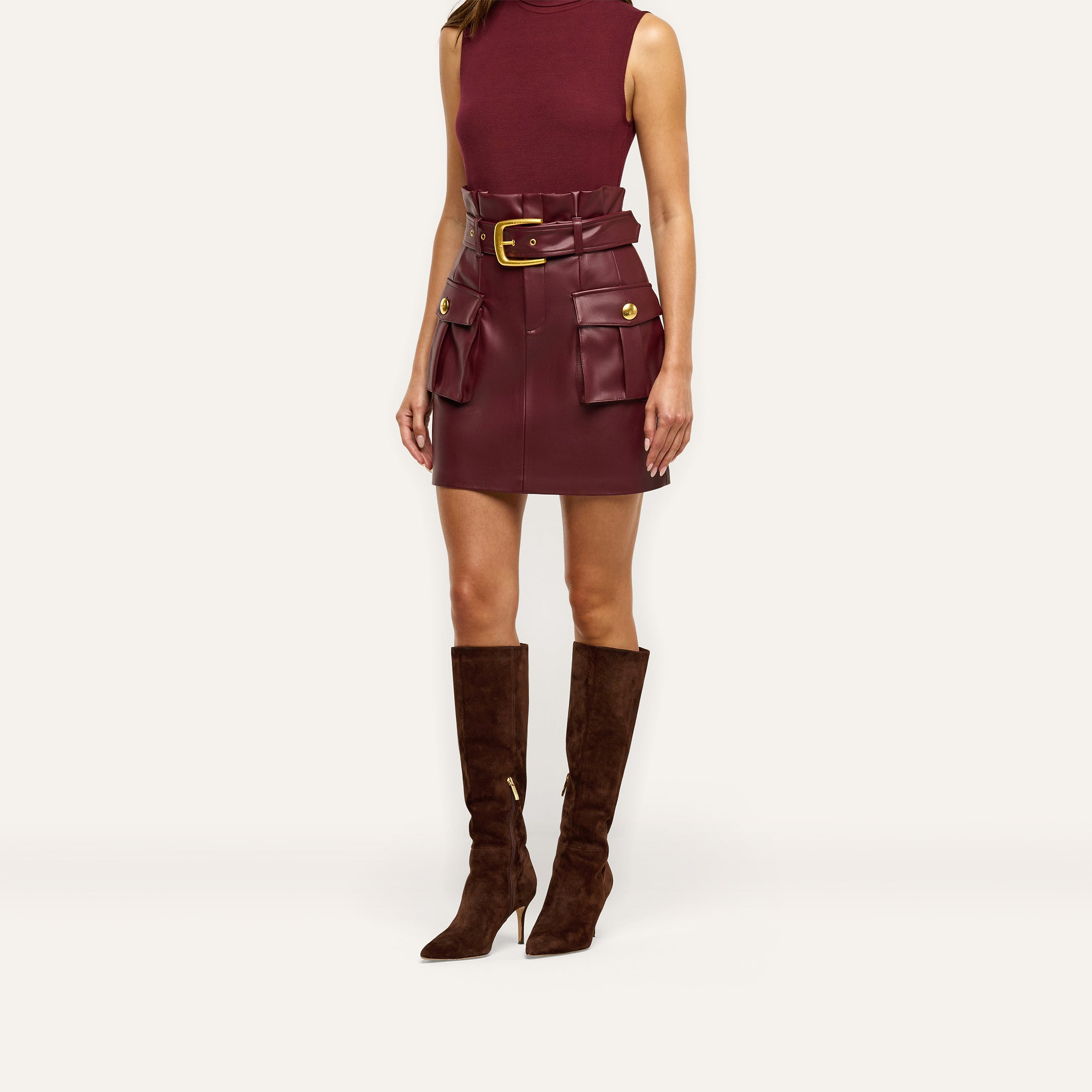 A burgundy Maelis Vegan Leather Skirt with a paperbag waist, large front cargo pockets, and a buckled belt, worn with a matching sleeveless top and brown knee-high boots.