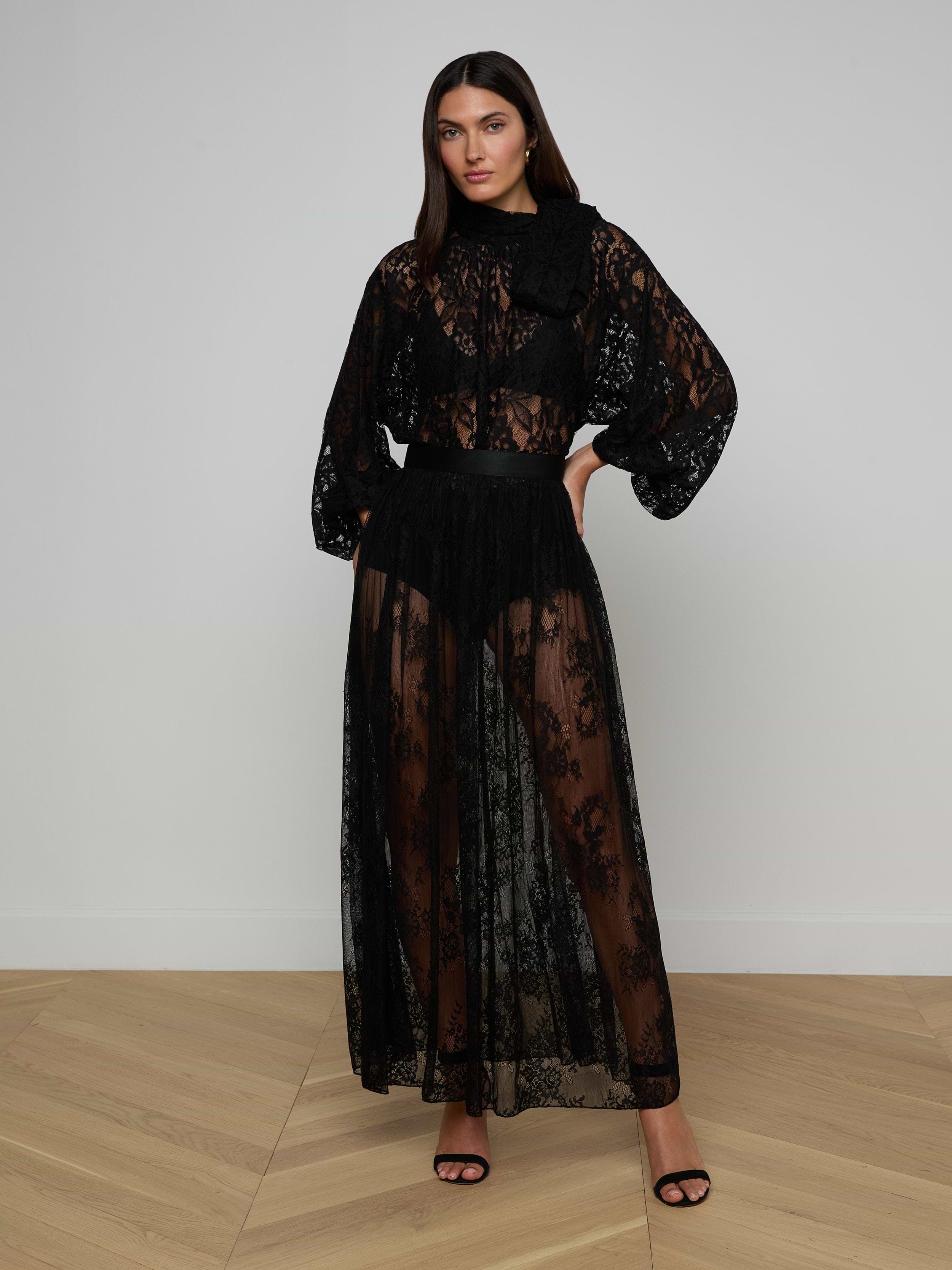 A woman stands against a plain white background wearing the Alona Lace Maxi Skirt in sheer black Chantilly floral lace, paired with a high-neck top featuring puffy sleeves and black high-heeled sandals.