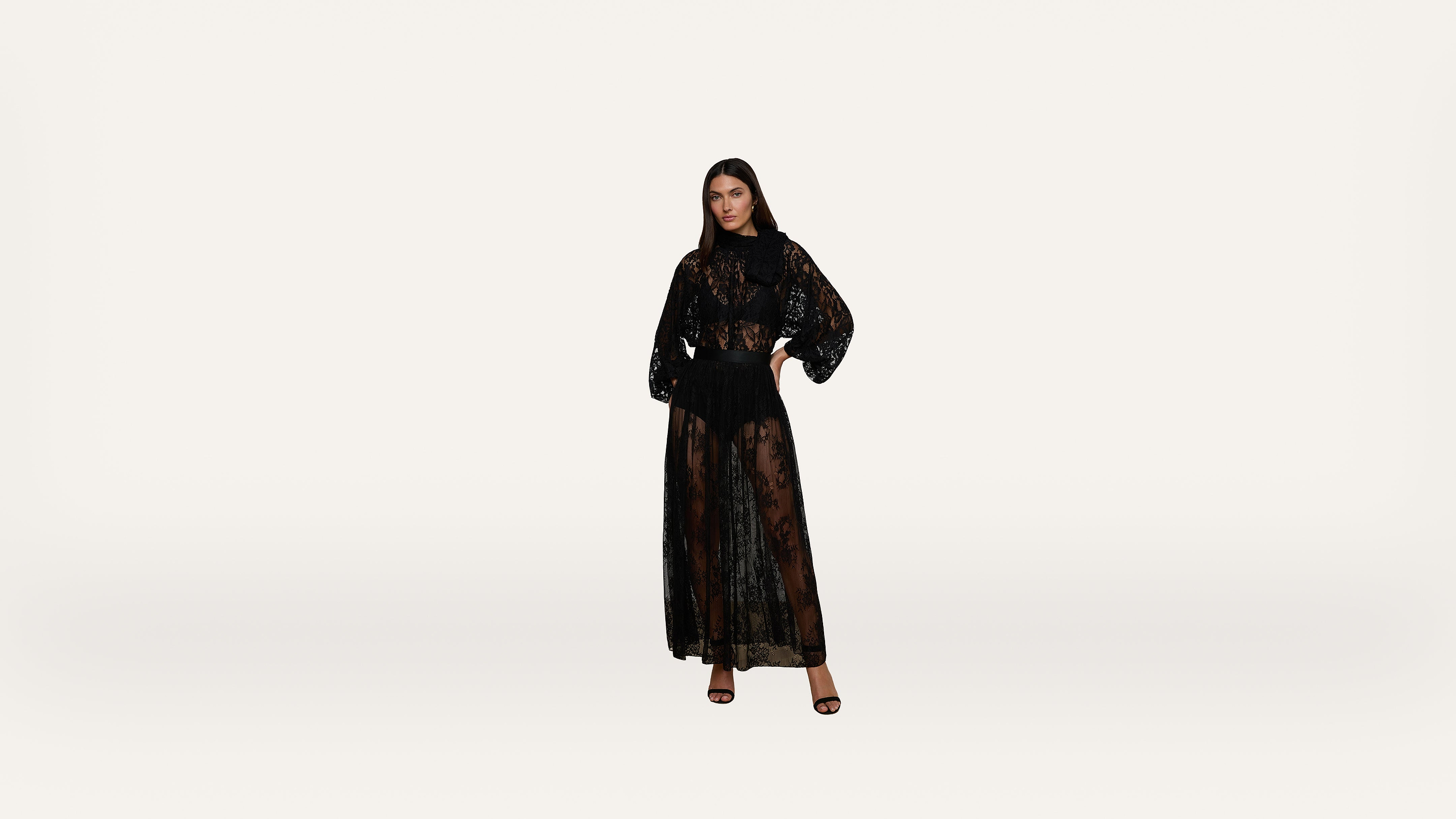 A woman stands against a plain white background, wearing a long, sheer black lace dress with a matching black undergarment and black high-heeled sandals.