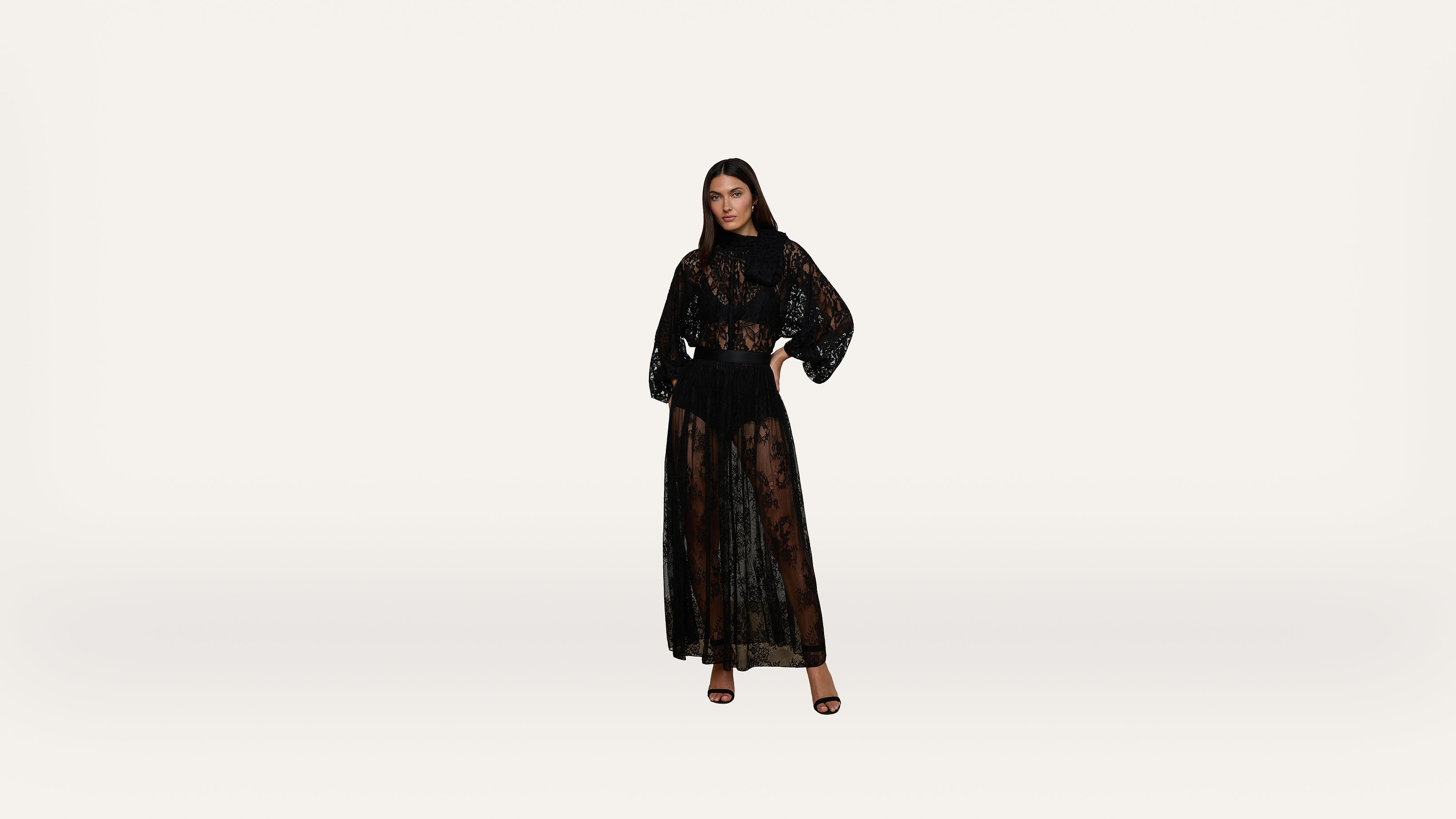 A woman stands against a plain white background, wearing a long, sheer black lace dress with a matching black undergarment and black high-heeled sandals.