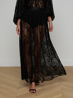 Wearing the Alona Lace Maxi Skirt in sheer Chantilly floral lace with a visible black undergarment and black heels, standing on a wooden floor against a plain wall—a bold, statement look for nights out.