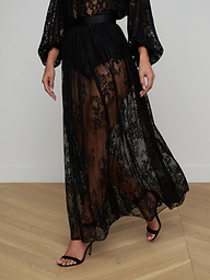 Wearing the Alona Lace Maxi Skirt—a long, sheer black Chantilly floral lace piece that delicately reveals the legs—paired with black high-heeled sandals, the person stands against a plain white background that heightens the sense of sheer romance.