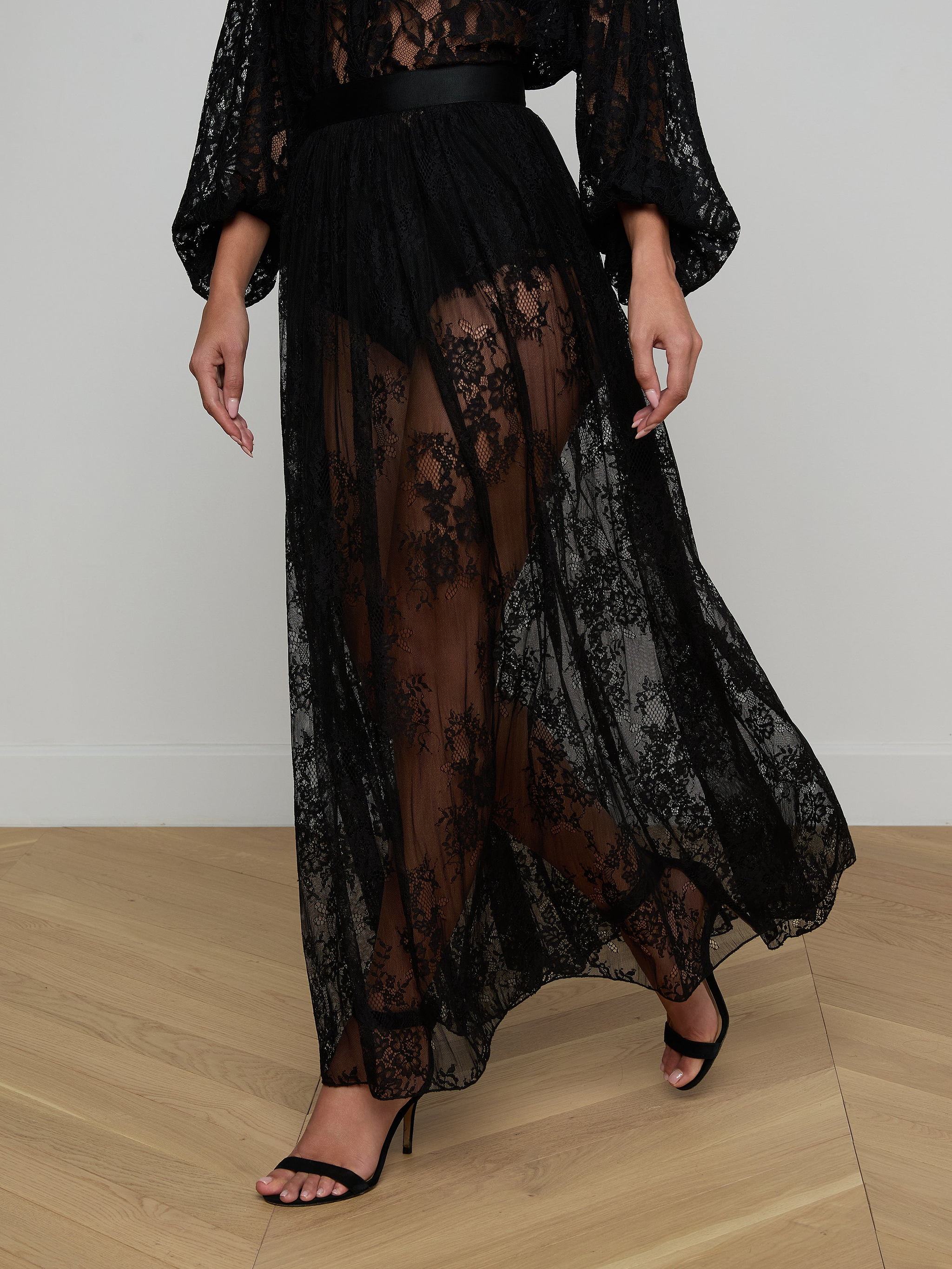Wearing the Alona Lace Maxi Skirt—a long, sheer black Chantilly floral lace piece that delicately reveals the legs—paired with black high-heeled sandals, the person stands against a plain white background that heightens the sense of sheer romance.