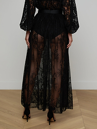 A person stands facing away, embodying sheer romance against a plain white background while wearing the Alona Lace Maxi Skirt in long, sheer black Chantilly floral lace paired with black high heels.