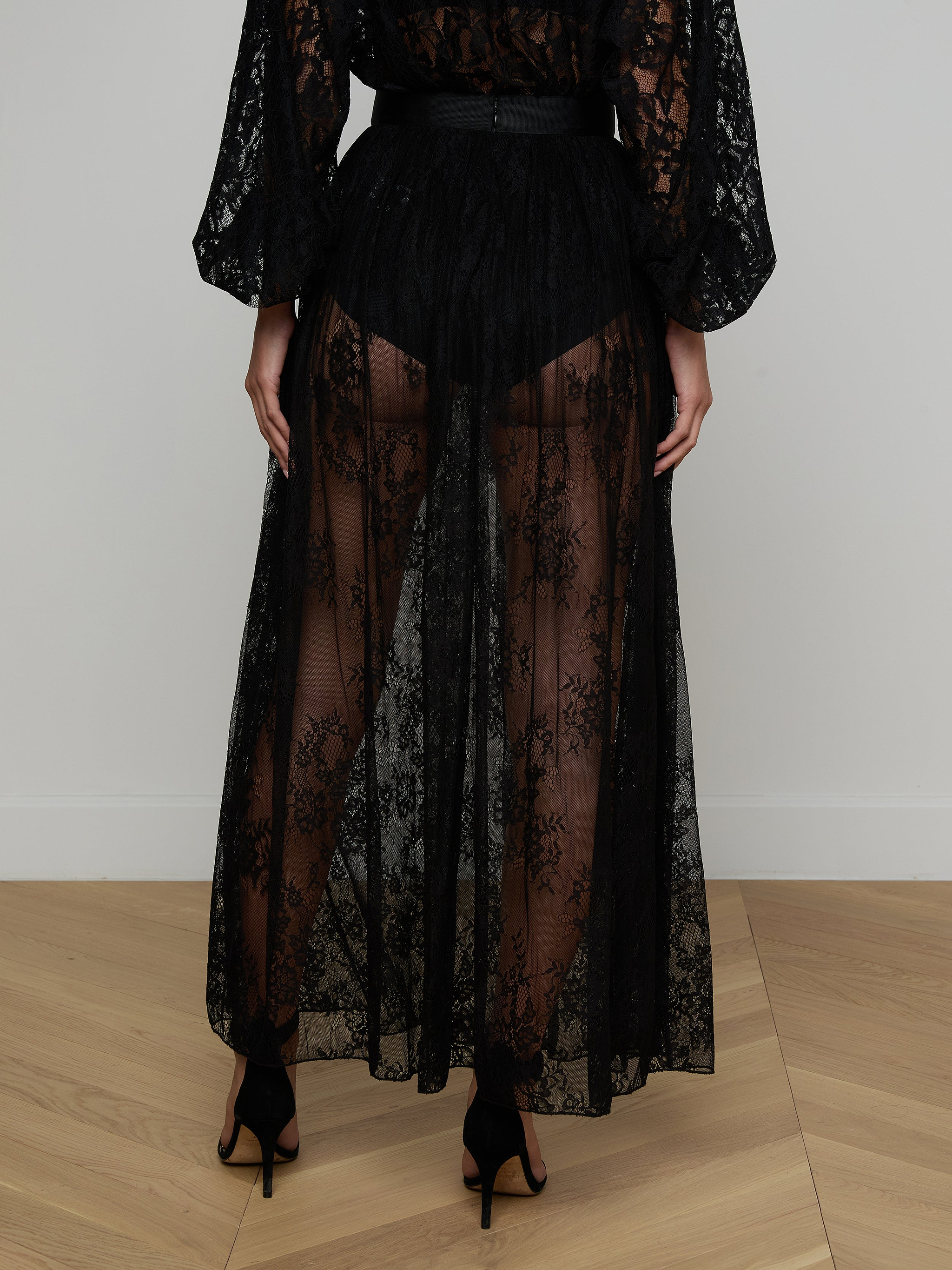 A person stands facing away, embodying sheer romance against a plain white background while wearing the Alona Lace Maxi Skirt in long, sheer black Chantilly floral lace paired with black high heels.