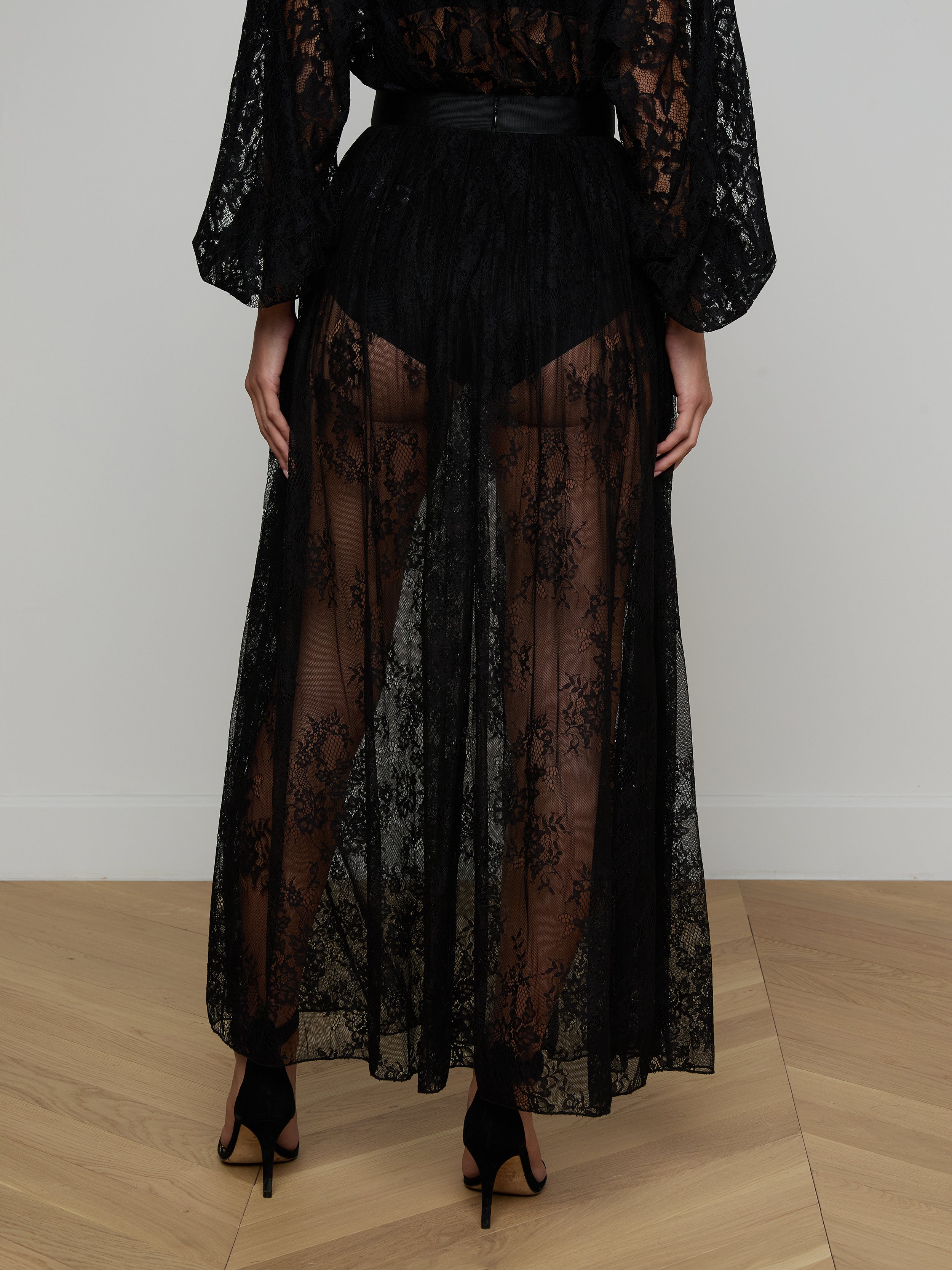 A person stands facing away, embodying sheer romance against a plain white background while wearing the Alona Lace Maxi Skirt in long, sheer black Chantilly floral lace paired with black high heels.