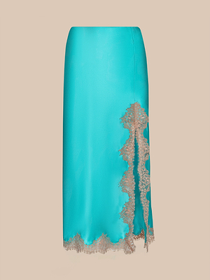 The Tico Silk Lace Skirt is a vibrant blue midi with beige eyelash lace trim and an asymmetrical right slit, shown on a plain beige background.