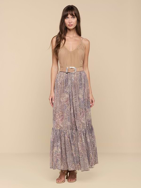 A woman stands facing forward against a plain white background, wearing the Ursie Maxi Skirt—a long, flowy bohemian style featuring a paisley leopard print—paired with a tan sleeveless top tucked in, cinched at the waist with a belt, and styled with brown sandals.