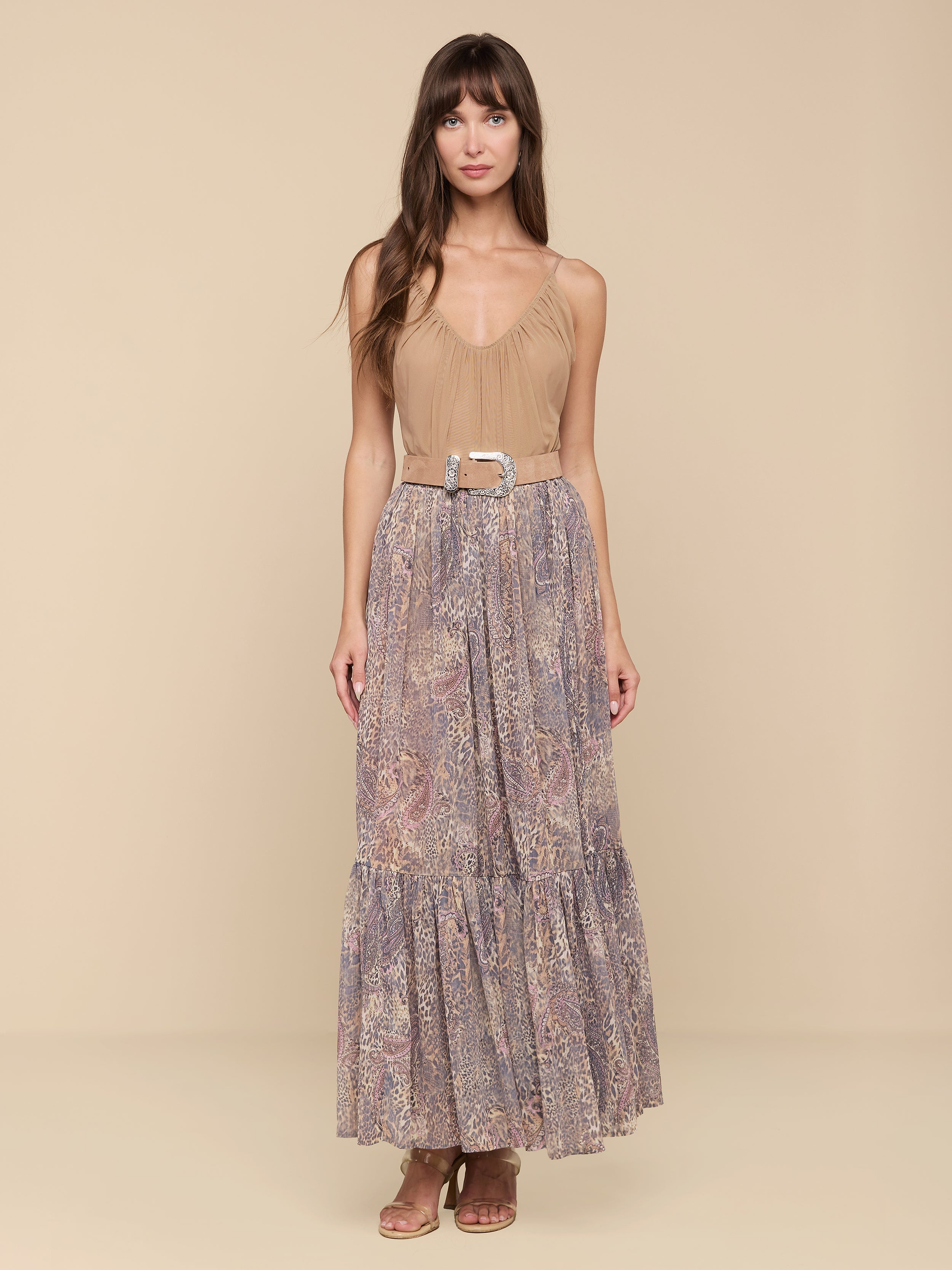 A woman stands facing forward against a plain white background, wearing the Ursie Maxi Skirt—a long, flowy bohemian style featuring a paisley leopard print—paired with a tan sleeveless top tucked in, cinched at the waist with a belt, and styled with brown sandals.
