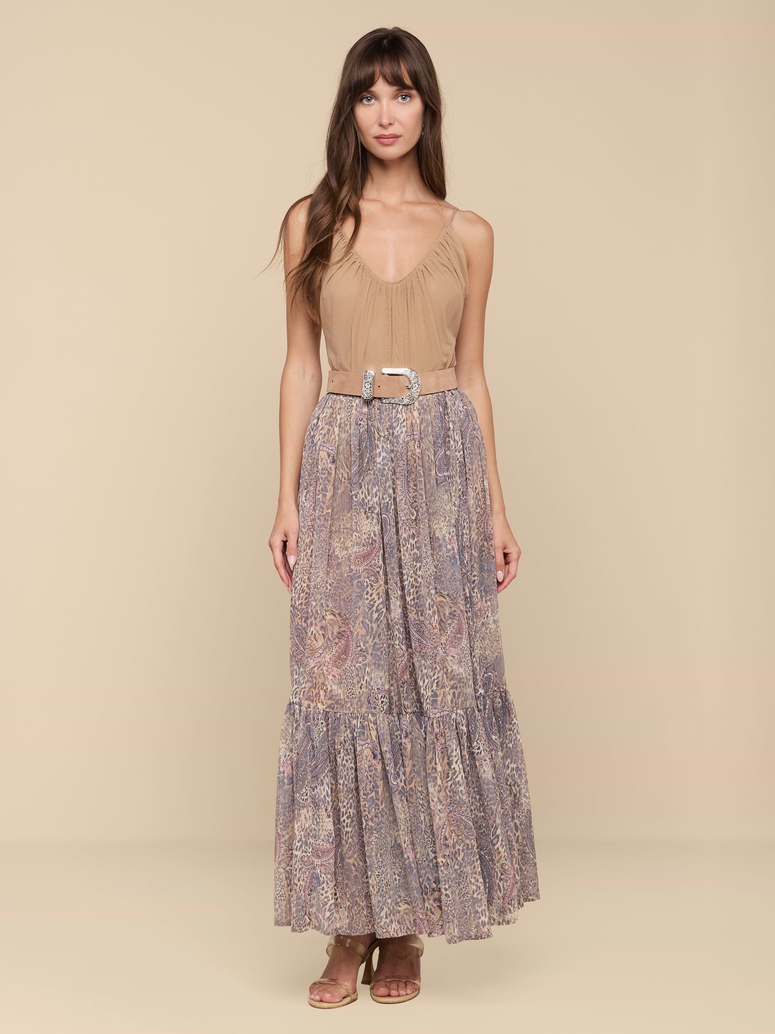 A woman stands facing forward against a plain white background, wearing the Ursie Maxi Skirt—a long, flowy bohemian style featuring a paisley leopard print—paired with a tan sleeveless top tucked in, cinched at the waist with a belt, and styled with brown sandals.