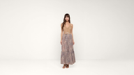Woman standing in a studio wearing a tan sleeveless top, a long floral skirt with a belt, and brown sandals, against a plain white background.