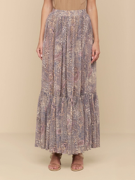 Wearing a tan top and the Ursie Maxi Skirt—an elegant, flowy bohemian skirt with ruffles at the hem and a paisley leopard print—a person pairs the look with flat sandals and stands against a plain white background.