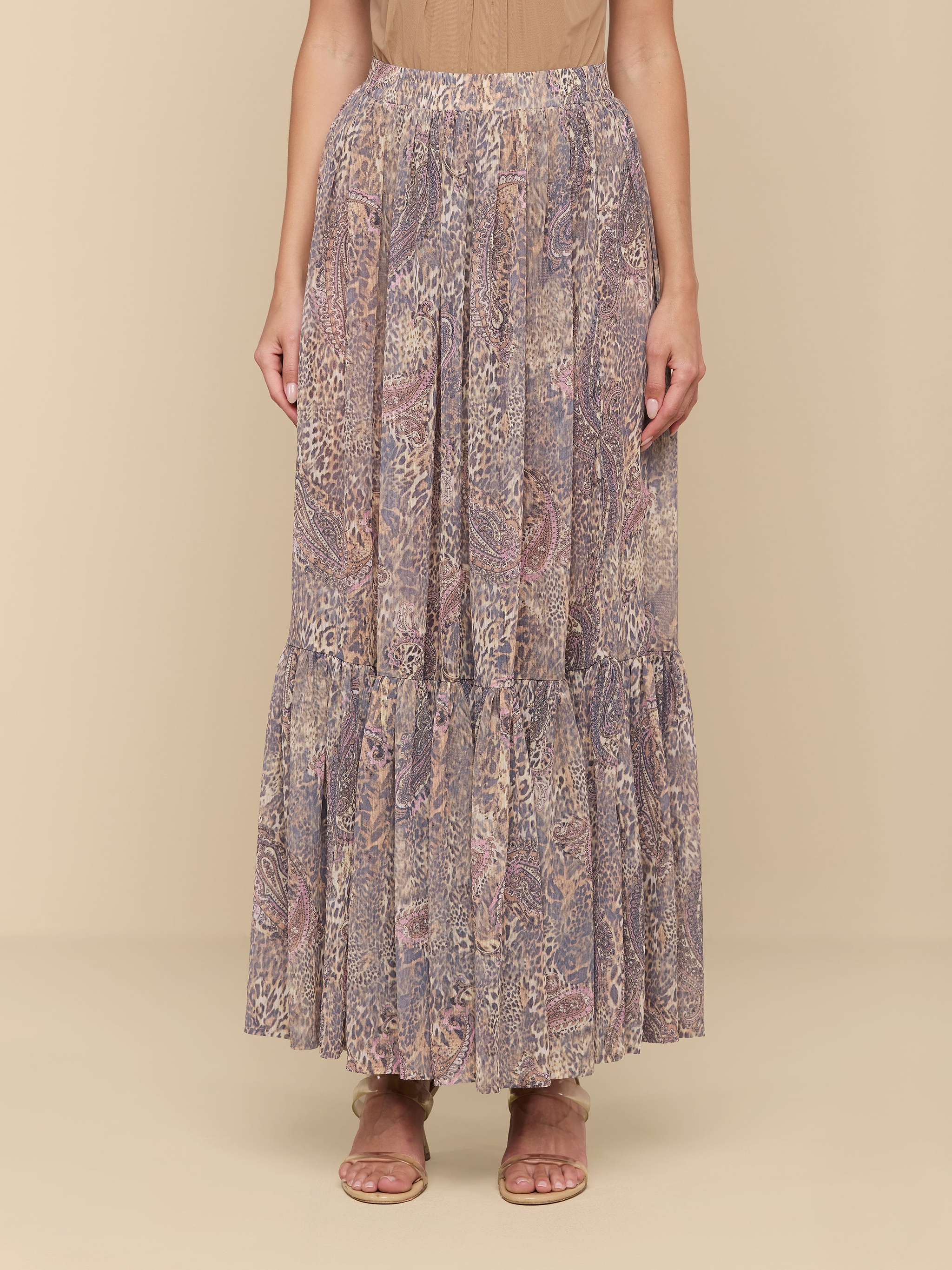Wearing a tan top and the Ursie Maxi Skirt—an elegant, flowy bohemian skirt with ruffles at the hem and a paisley leopard print—a person pairs the look with flat sandals and stands against a plain white background.
