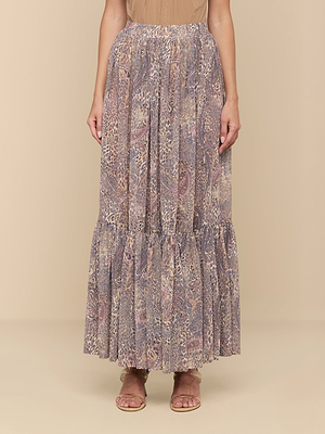 A person wearing the Ursie Maxi Skirt—a long, flowing skirt with a muted animal print in purple, beige, and brown with a ruffled hem—stands against a plain beige background, paired with sandals.