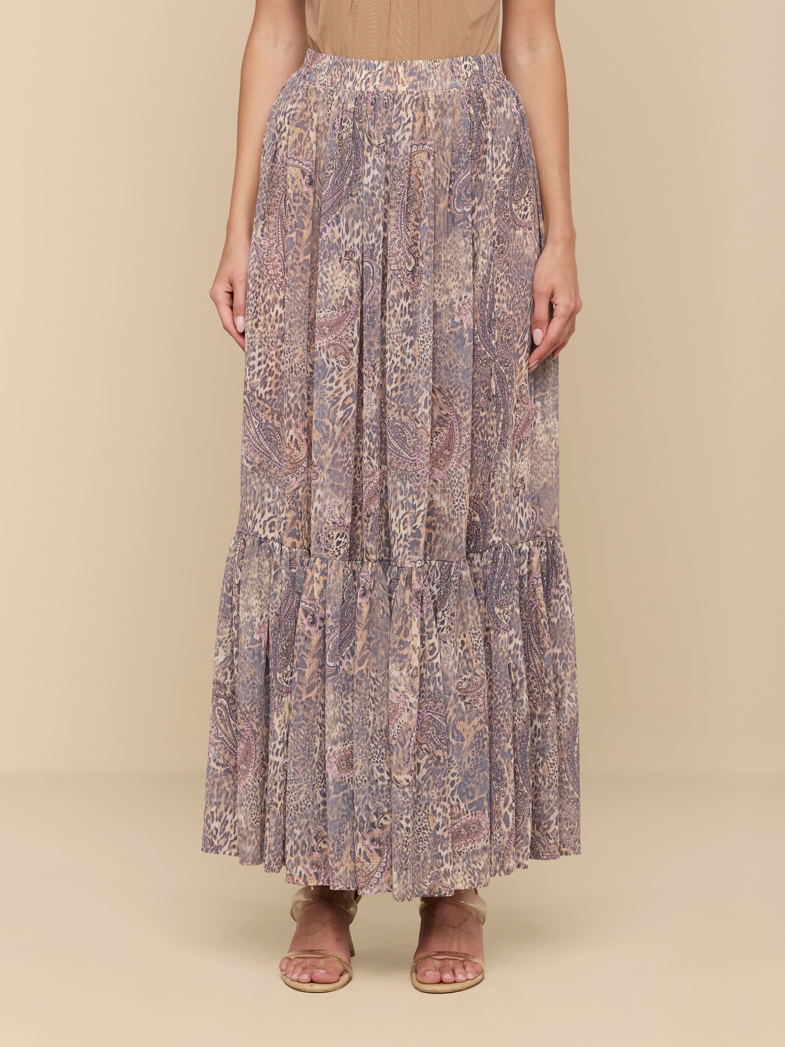 Wearing a tan top and the Ursie Maxi Skirt—an elegant, flowy bohemian skirt with ruffles at the hem and a paisley leopard print—a person pairs the look with flat sandals and stands against a plain white background.