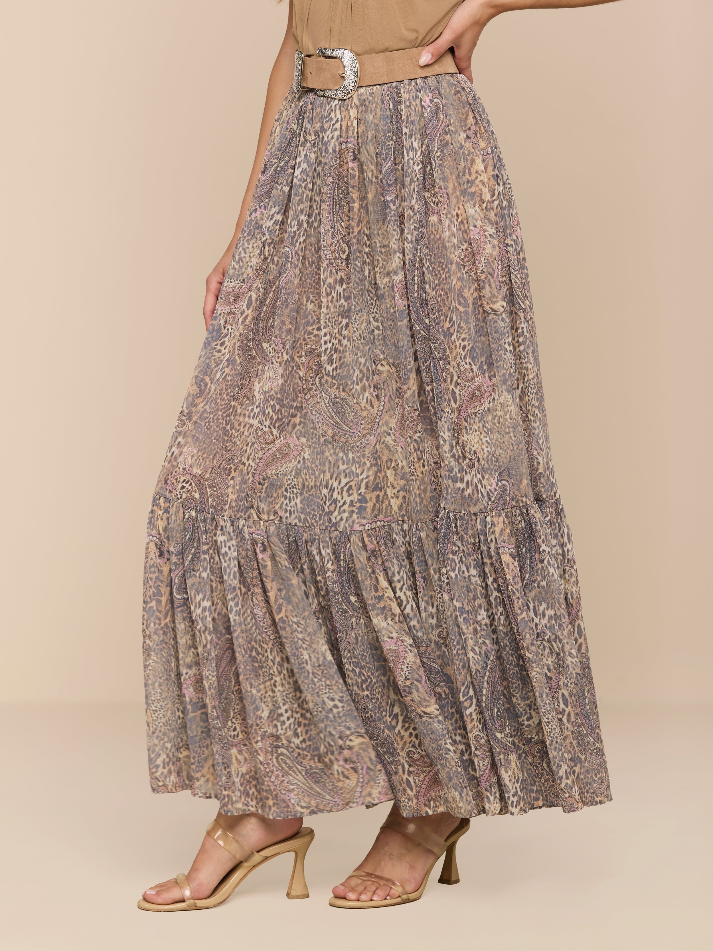 A woman models the Ursie Maxi Skirt—a flowy, ankle-length paisley leopard print design with a belt—paired with a beige top and tan heeled sandals against a plain white background. Her face is not visible in the image.