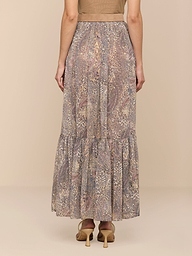 A woman stands facing away, wearing a brown top and the Ursie Maxi Skirt—a long, bohemian skirt with a paisley leopard print and a tiered ruffle at the hem—paired with tan high-heeled shoes against a plain white background.