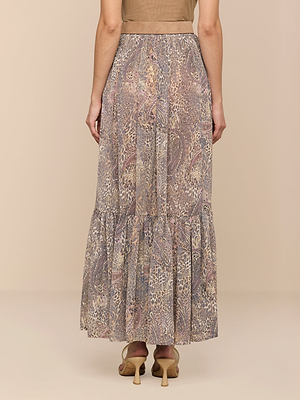 A woman is seen from the back wearing the Ursie Maxi Skirt, featuring a muted paisley and animal print. She pairs it with light heeled sandals and a beige top. The background is plain and neutral.