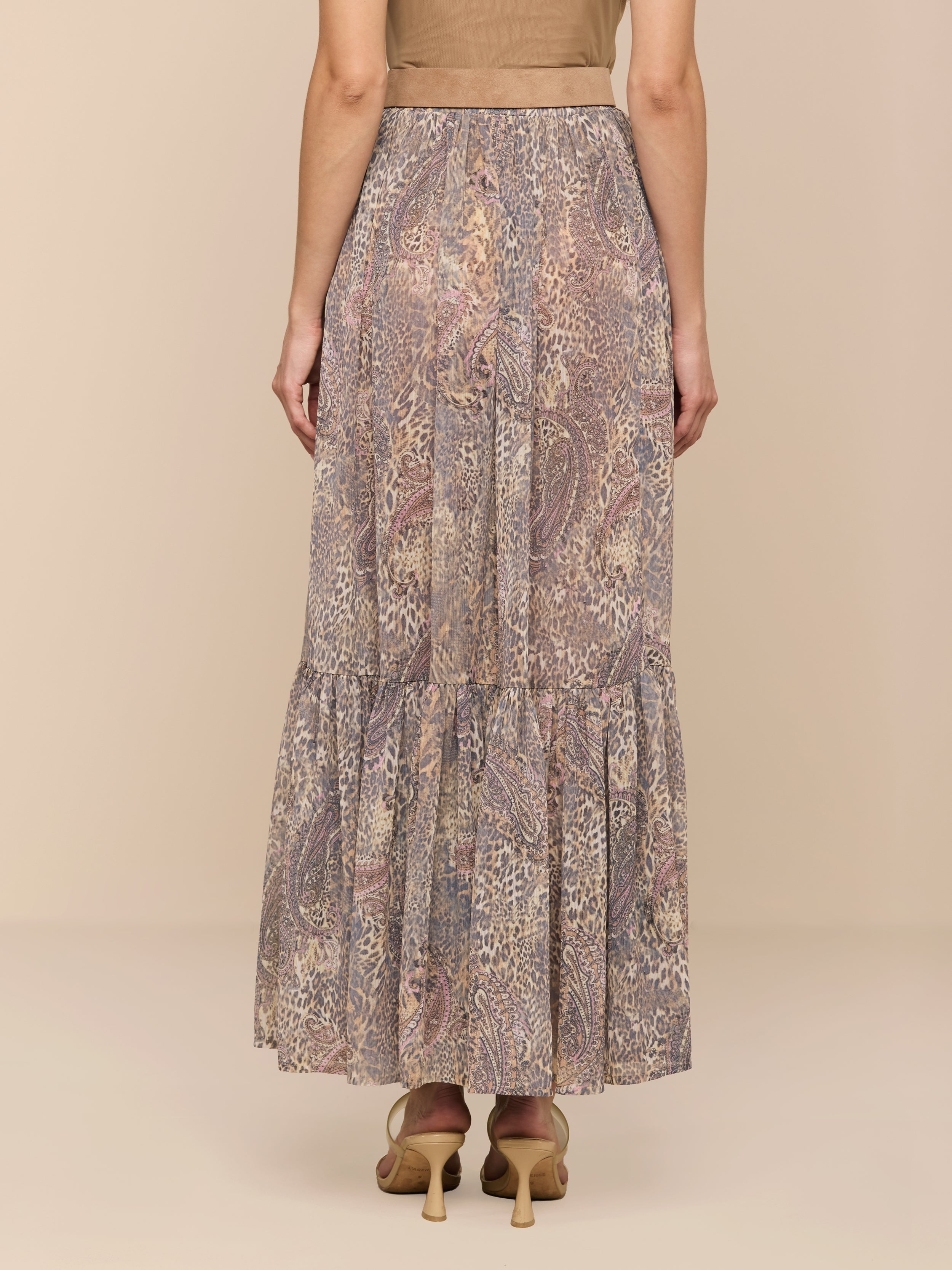 A woman stands facing away, wearing a brown top and the Ursie Maxi Skirt—a long, bohemian skirt with a paisley leopard print and a tiered ruffle at the hem—paired with tan high-heeled shoes against a plain white background.