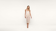 Woman standing against a white background, wearing a sleeveless white top, a beige lace midi skirt with buttons, and beige high-heeled sandals. She has long, blonde hair and is smiling at the camera.