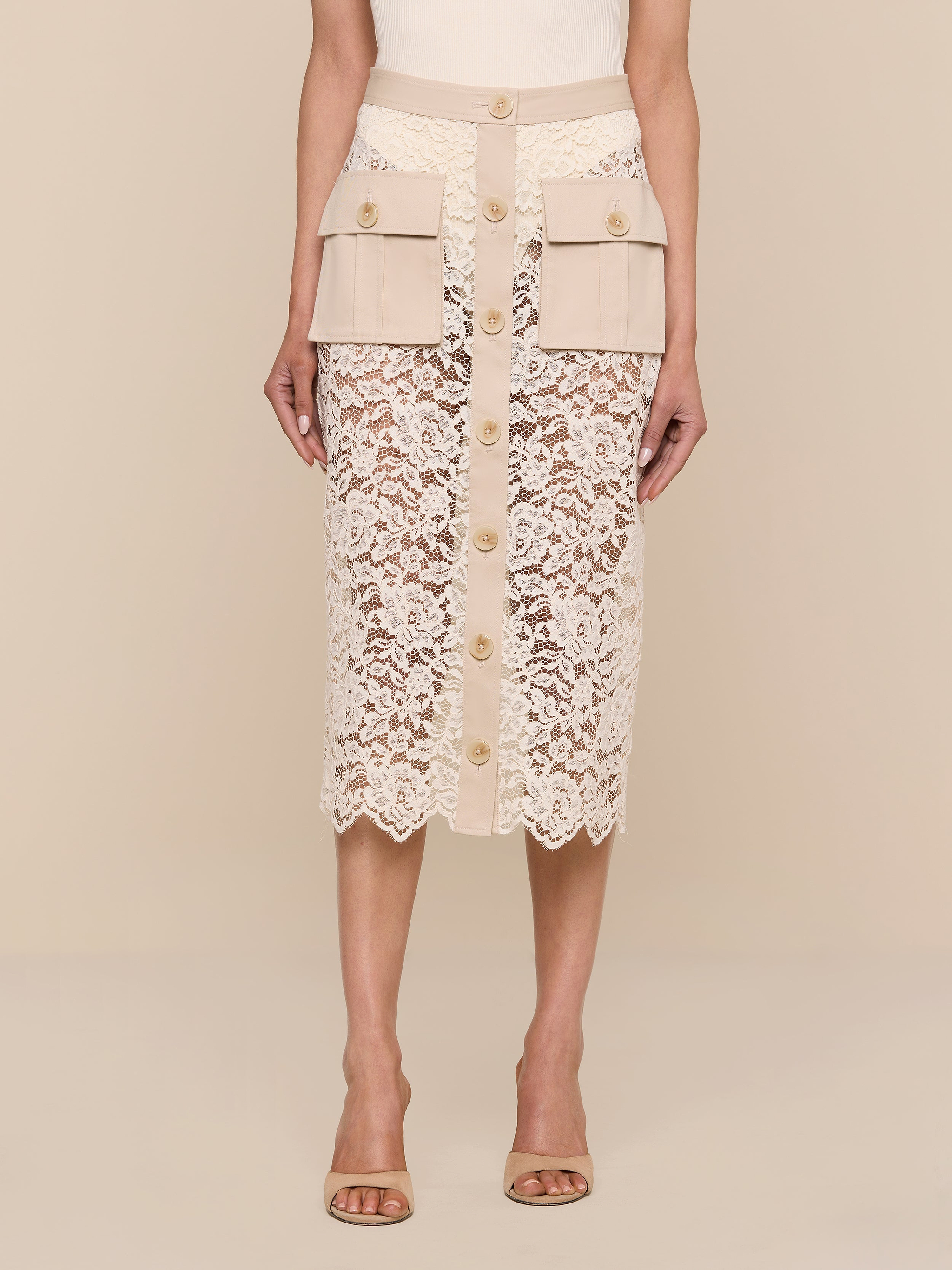 A woman stands against a plain white background wearing the Shae Skirt—a beige floral lace midi skirt featuring a button-down front and cargo pockets—paired with nude heeled sandals.