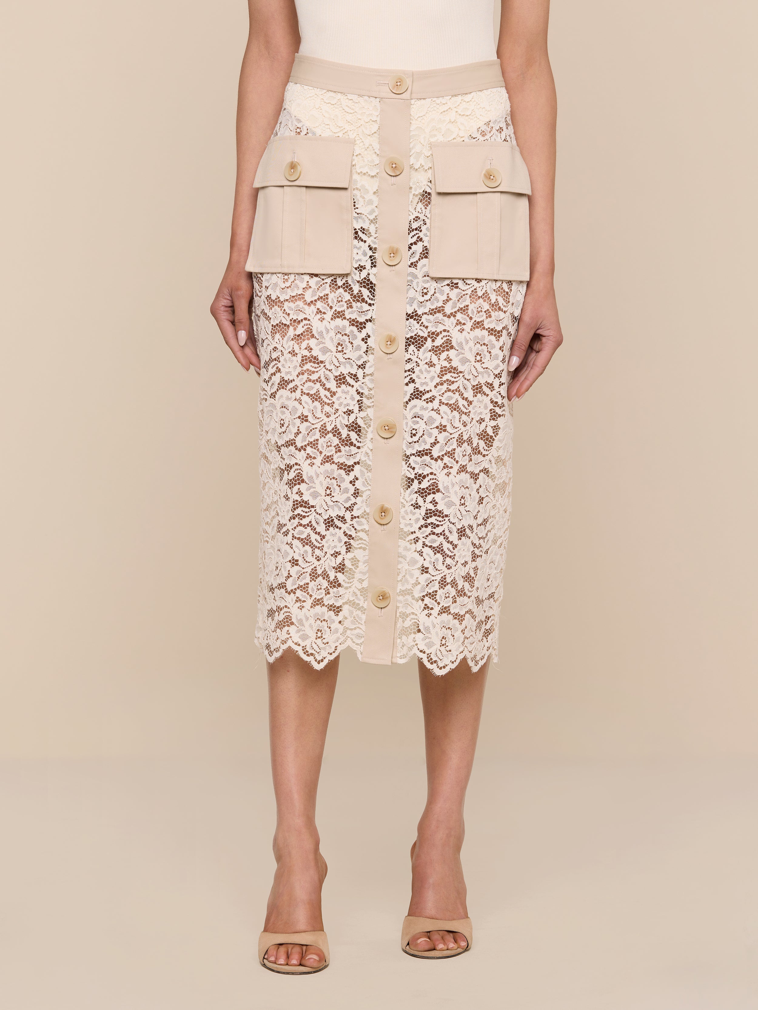 A woman stands against a plain white background wearing the Shae Skirt—a beige floral lace midi skirt featuring a button-down front and cargo pockets—paired with nude heeled sandals.