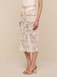 A woman wearing the Shae Skirt—a beige floral lace midi skirt with large cargo pockets, a high waist, and button-down detail—paired with a white top and beige heeled sandals, stands against a plain white background.