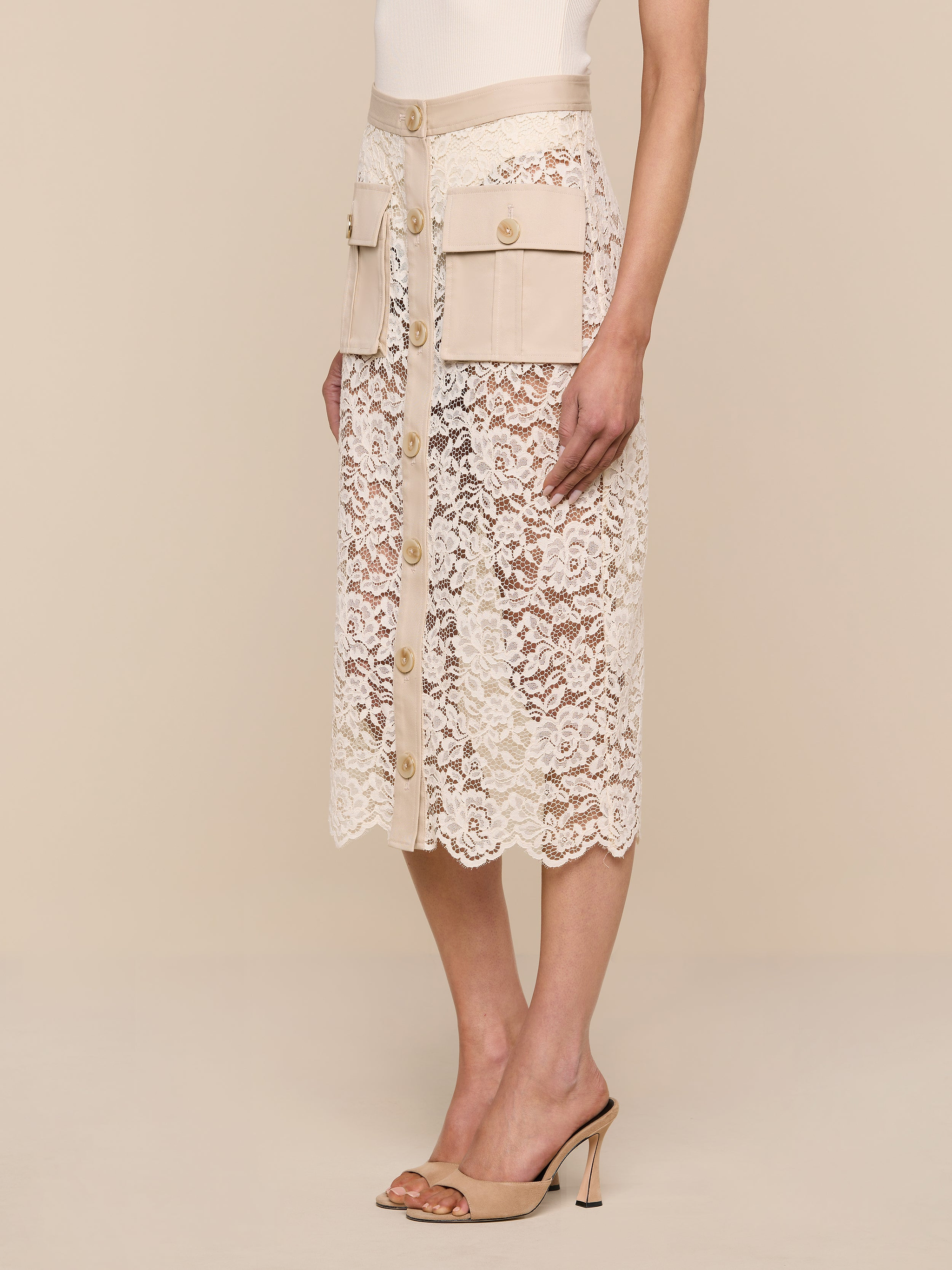 A woman wearing the Shae Skirt—a beige floral lace midi skirt with large cargo pockets, a high waist, and button-down detail—paired with a white top and beige heeled sandals, stands against a plain white background.