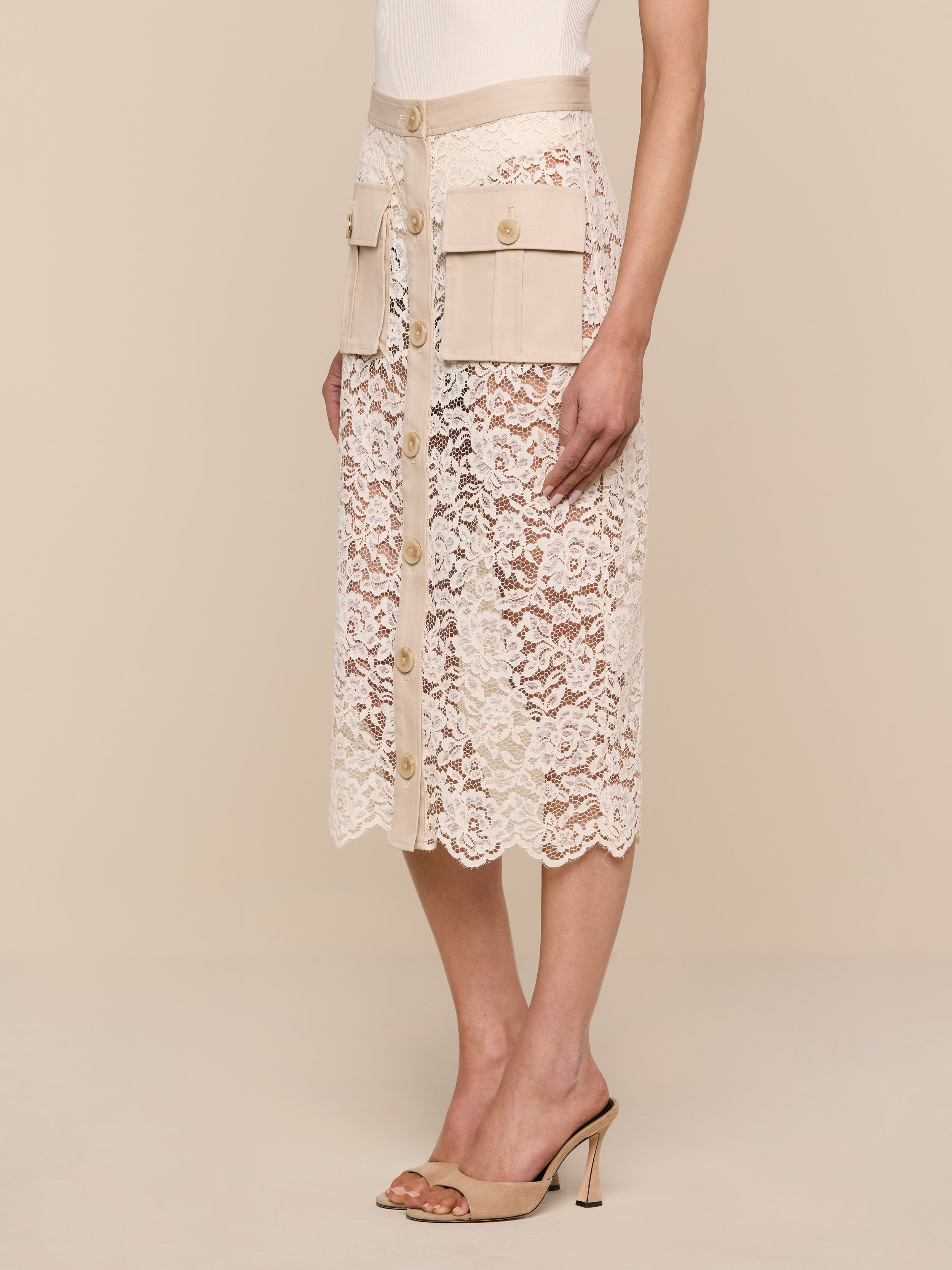 A woman wearing the Shae Skirt—a beige floral lace midi skirt with large cargo pockets, a high waist, and button-down detail—paired with a white top and beige heeled sandals, stands against a plain white background.