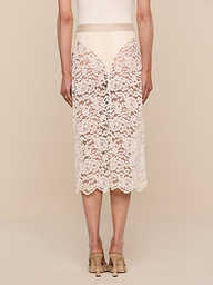 A person wearing the Shae Skirt—a knee-length, beige floral lace skirt—and beige heels is standing with their back to the camera against a plain white background.