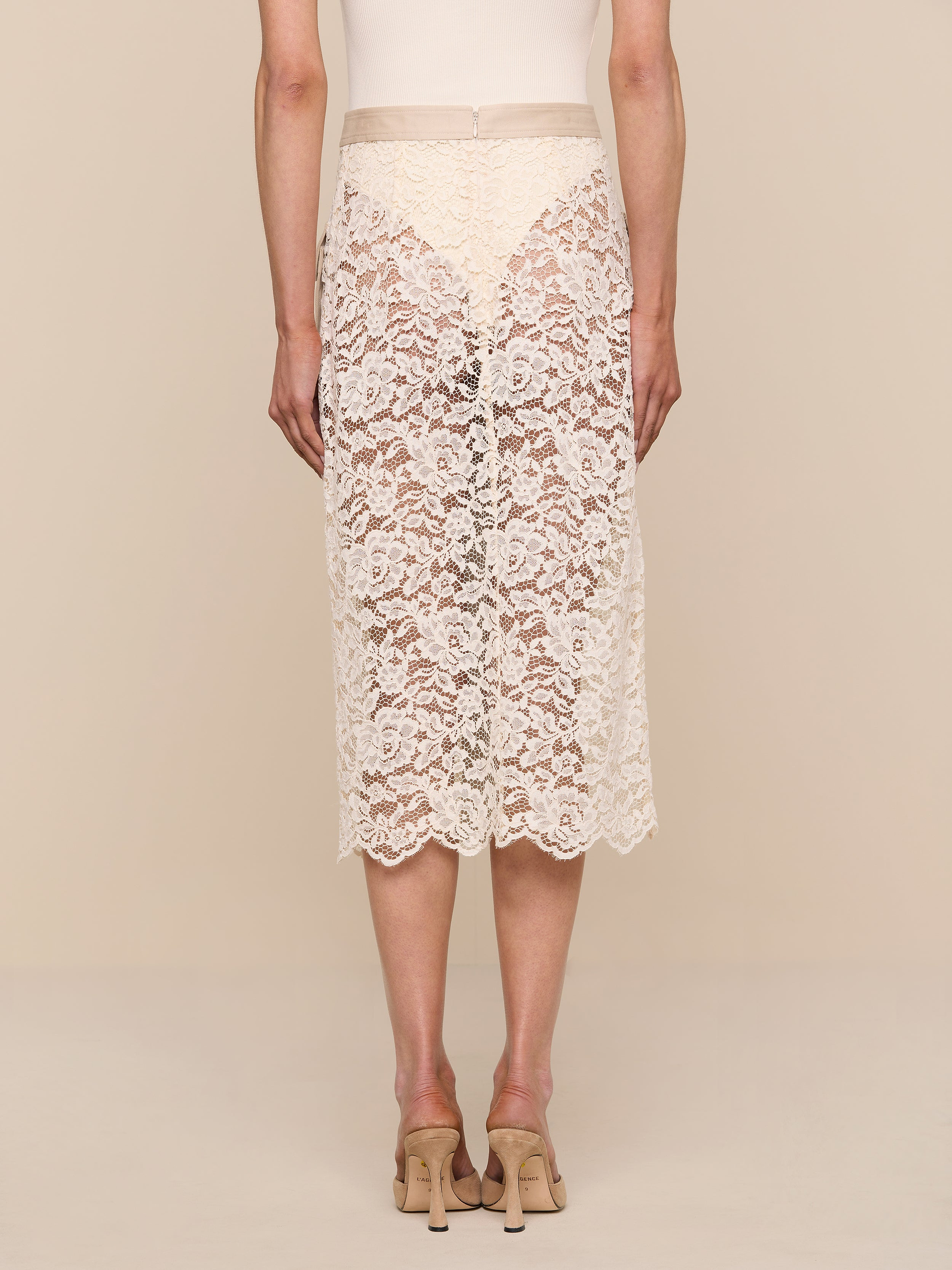 A person wearing the Shae Skirt—a knee-length, beige floral lace skirt—and beige heels is standing with their back to the camera against a plain white background.