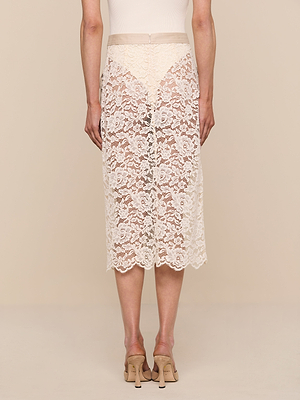 A person stands facing away, wearing a beige top, the Shae Skirt—a sheer floral lace midi—and beige high-heeled sandals against a neutral background.