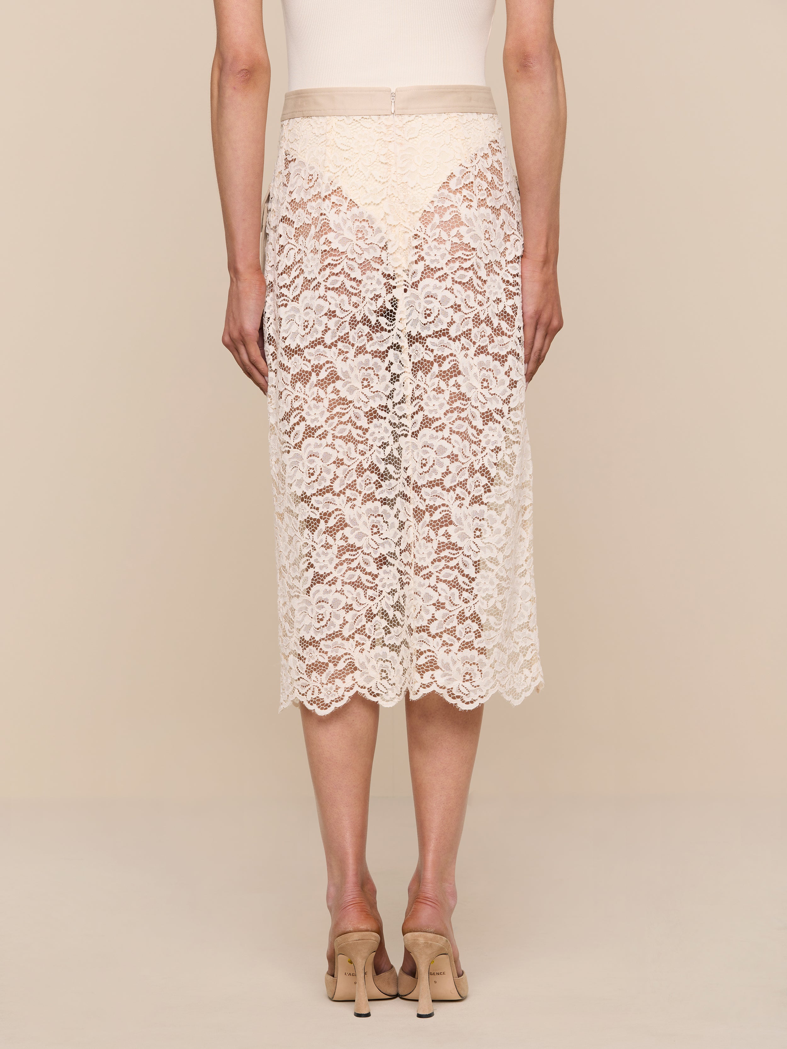 A person wearing the Shae Skirt—a knee-length, beige floral lace skirt—and beige heels is standing with their back to the camera against a plain white background.