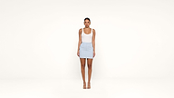 A woman stands against a plain white background, wearing a white tank top, a light blue mini skirt, and nude heeled sandals. She faces forward with her arms relaxed at her sides.