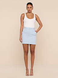 A woman stands facing forward, wearing a white tank top, the Vera Pinstriped Mini Skirt in a light blue cotton blend, and nude heeled sandals, set against a plain white background.