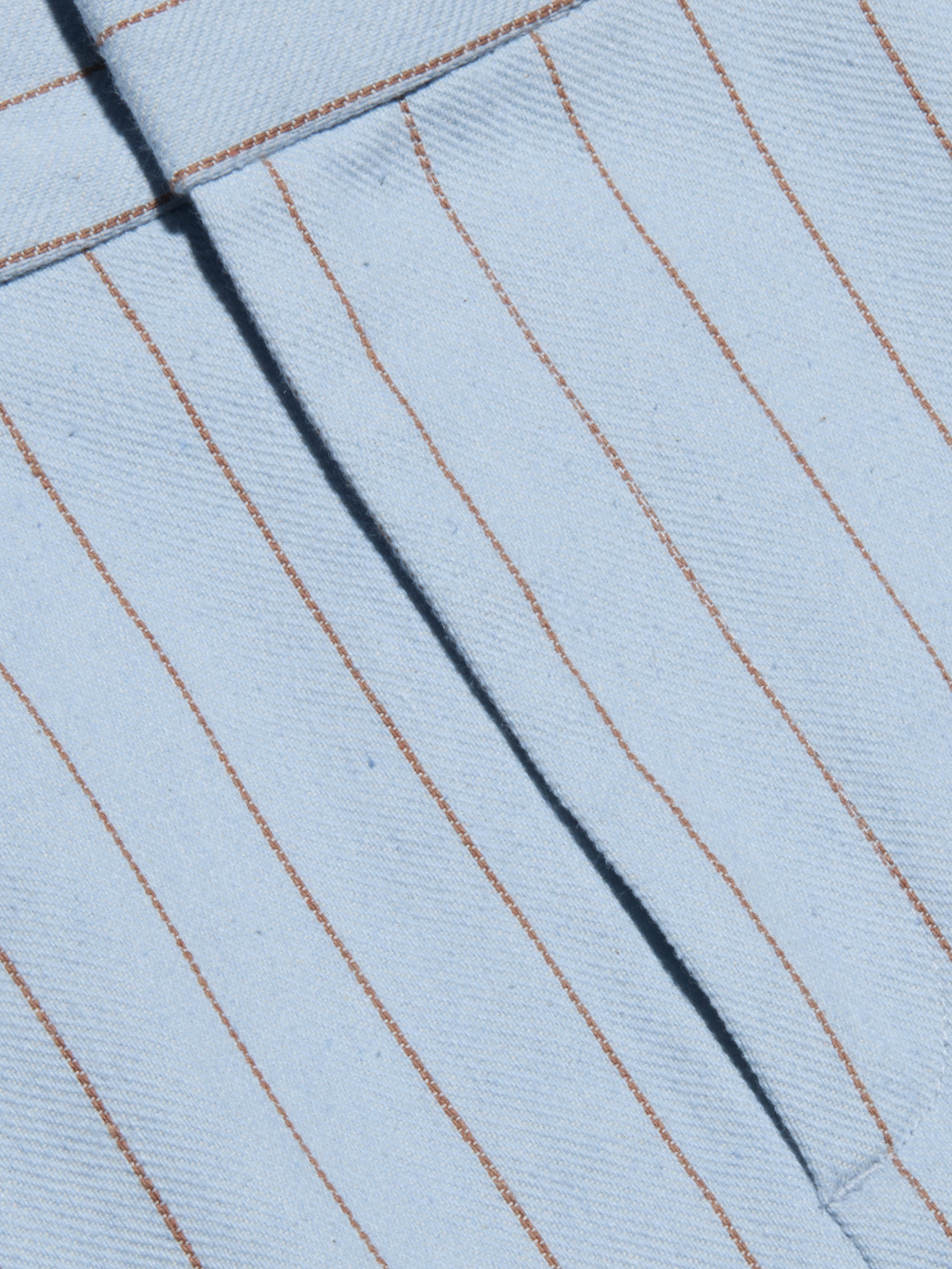 Close-up of the Vera Pinstriped Mini Skirt, crafted from a light blue cotton blend fabric featuring thin, evenly spaced vertical brown pinstripes and a visible seam running diagonally across the tailored silhouette.