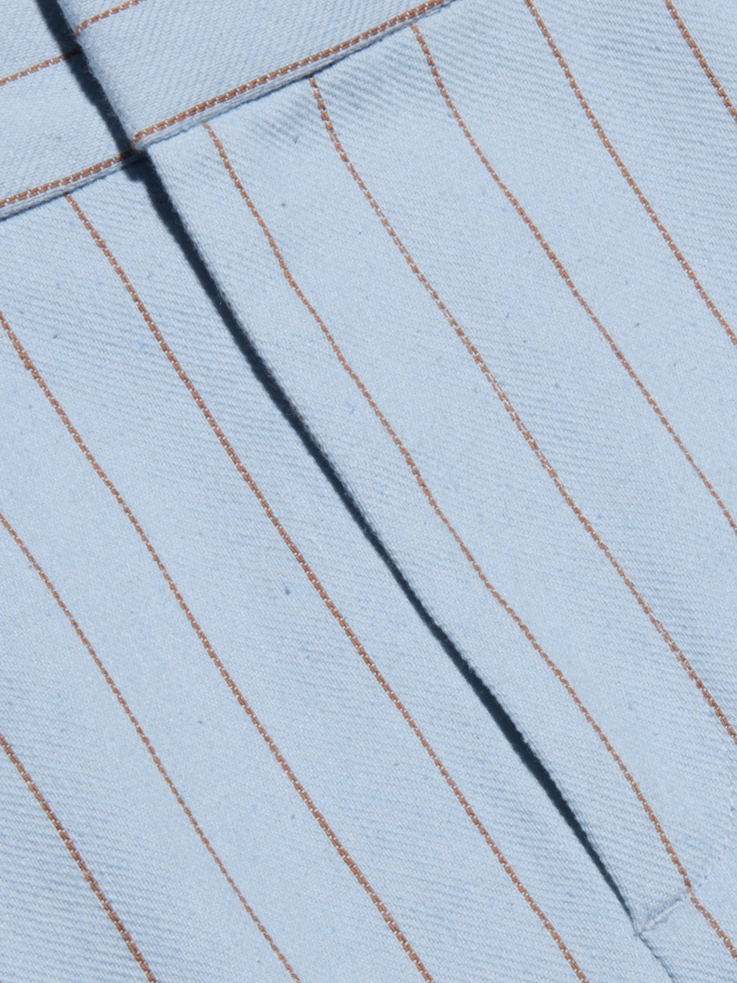 Close-up of the Vera Pinstriped Mini Skirt, crafted from a light blue cotton blend fabric featuring thin, evenly spaced vertical brown pinstripes and a visible seam running diagonally across the tailored silhouette.