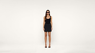 Woman wearing sunglasses, a black tank top, black skirt, and black heels stands against a plain white background. She faces forward with arms at her sides.