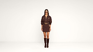 A woman stands against a plain white background, wearing a brown belted dress with pockets, knee-high brown boots, sunglasses, and gold accessories.