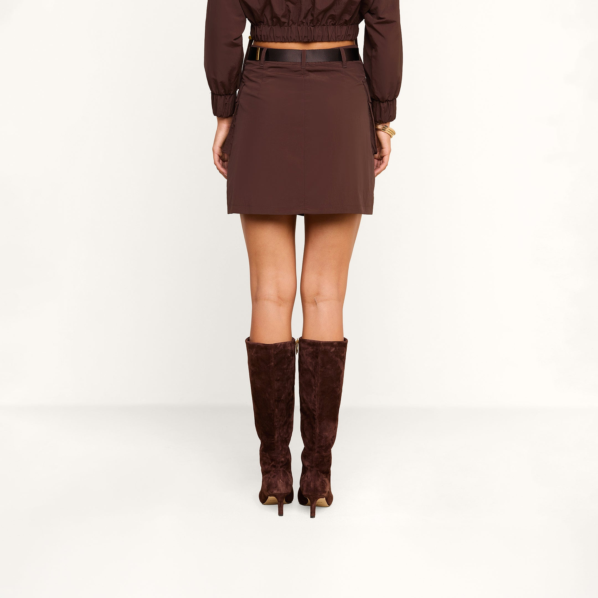 A person stands facing away against a plain white background, wearing the Norene Nylon Mini Skirt in brown, paired with matching knee-high suede boots and a cropped brown jacket.