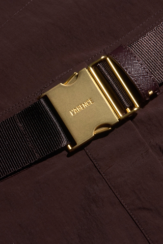 A close-up of a gold metal buckle engraved with the word PATIENTS, attached to a dark brown strap on the Norene Nylon Mini Skirt, which features water-repellent fabric, set against a dark brown textile background.