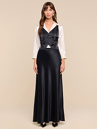 A woman with long brown hair and bangs stands facing forward against a plain white background, wearing the Vespera Suspender Maxi Skirt paired with a white blouse and a black vest.