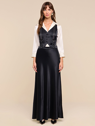 A woman stands against a plain background, dressed in a white collared blouse and black buttoned vest, paired with the Vespera Suspender Maxi Skirt. She completes her look with black shoes, while her long brown hair frames her neutral expression.