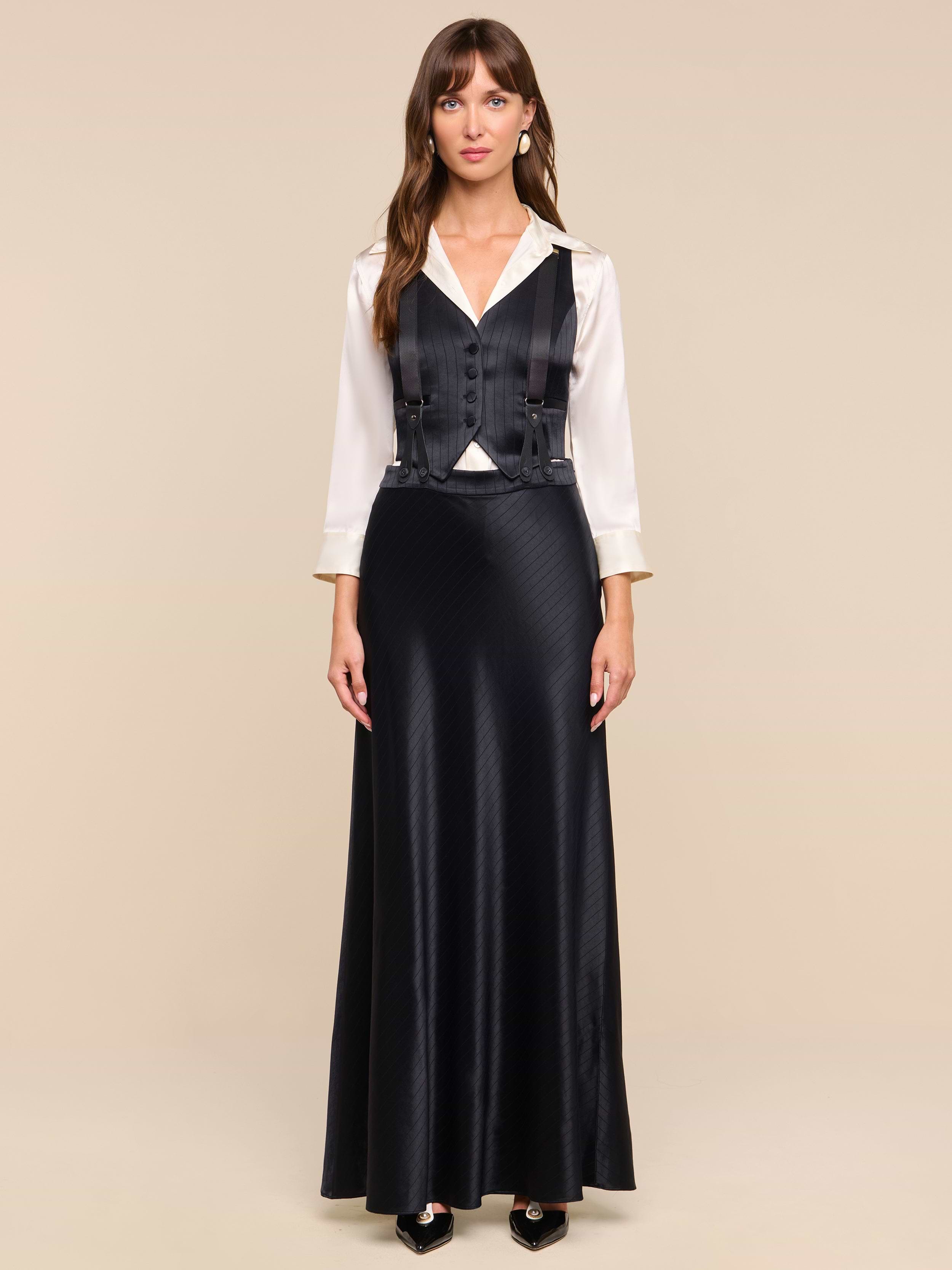 A woman with long brown hair and bangs stands facing forward against a plain white background, wearing the Vespera Suspender Maxi Skirt paired with a white blouse and a black vest.
