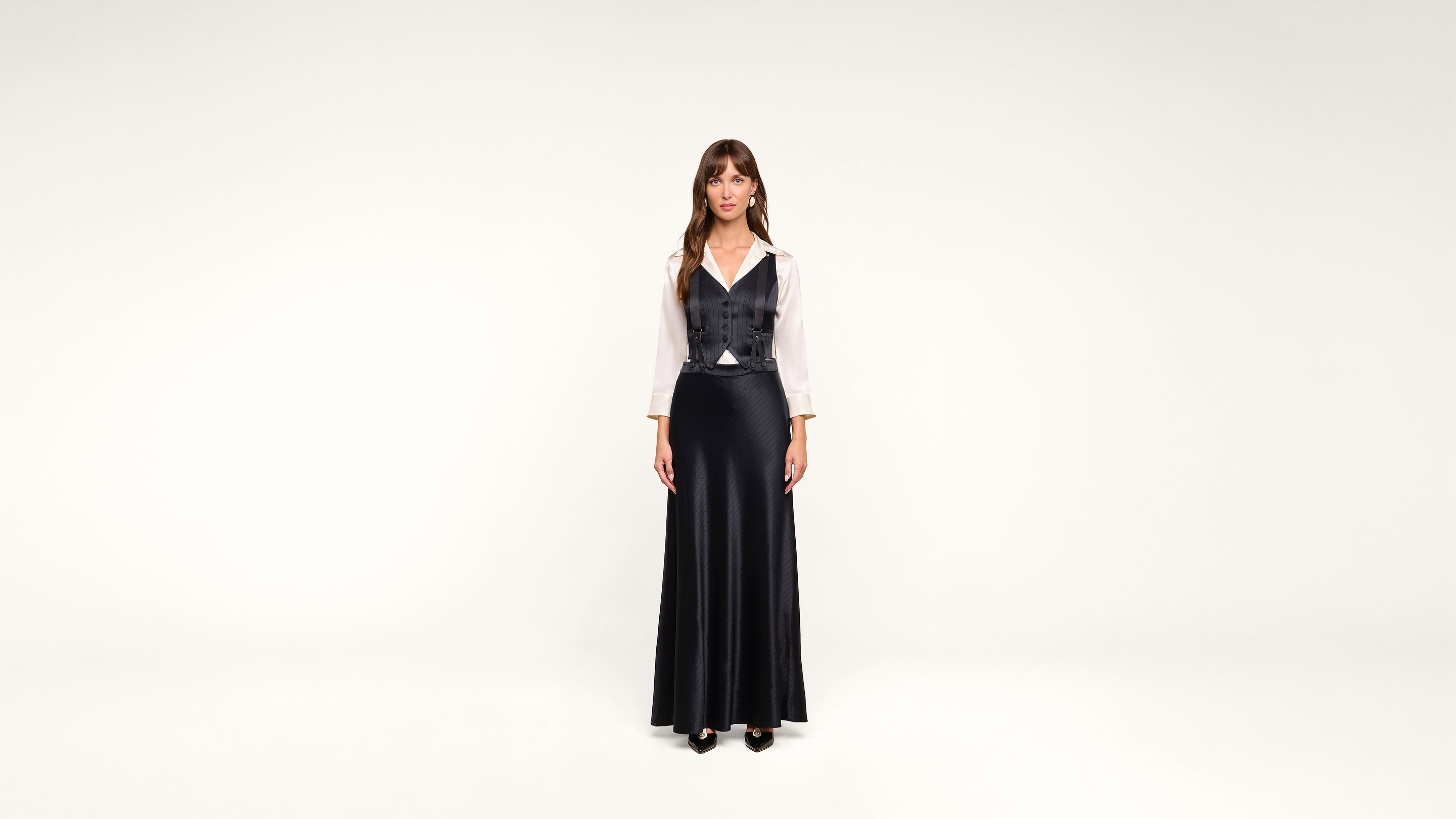 A woman stands against a plain white background, wearing a long black skirt, a white blouse, and a black vest. She faces forward with her arms at her sides and has long brown hair with bangs.
