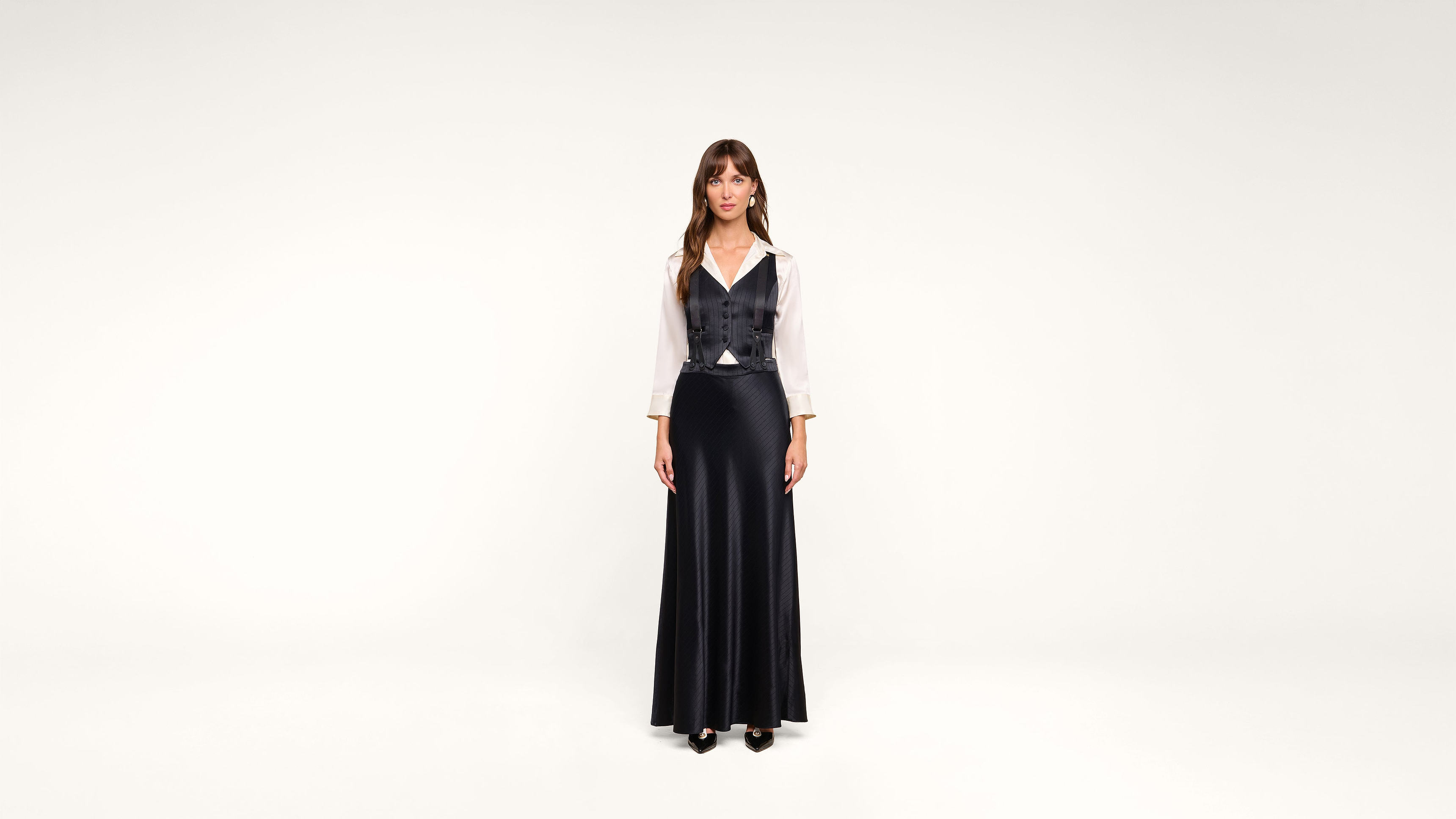 A woman stands against a plain white background, wearing a long black skirt, a white blouse, and a black vest. She faces forward with her arms at her sides and has long brown hair with bangs.