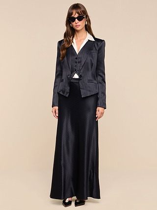 A woman wearing sunglasses, a dark pinstriped blazer, matching vest, adjustable suspenders over a white collared shirt, and the Vespera Suspender Maxi Skirt stands against a plain beige background.