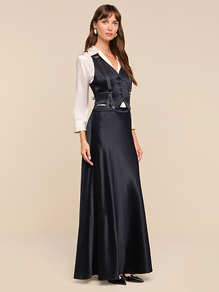 A woman stands wearing the Vespera Suspender Maxi Skirt, paired with a white blouse and a matching black vest featuring buttons and lapels. She has long brown hair, wears black pointed shoes, and poses against a plain beige background.