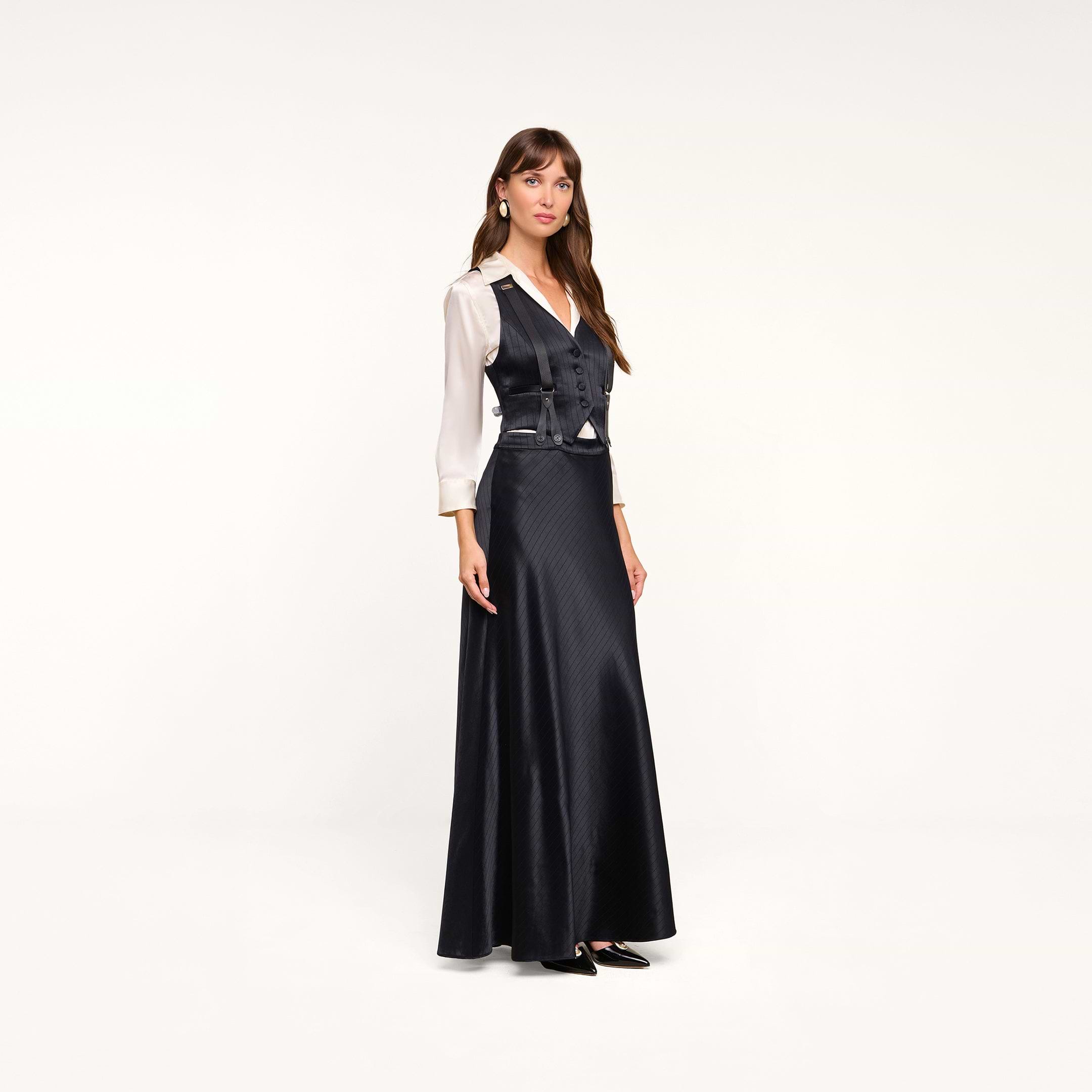 Wearing the Vespera Suspender Maxi Skirt, a woman poses against a plain white background, styling the long black pinstripe skirt with a fitted vest-style top over a white blouse.